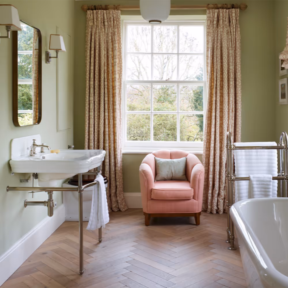 A period bathroom painted olive green with a blush pink armchair upon a engineered wooden floor.