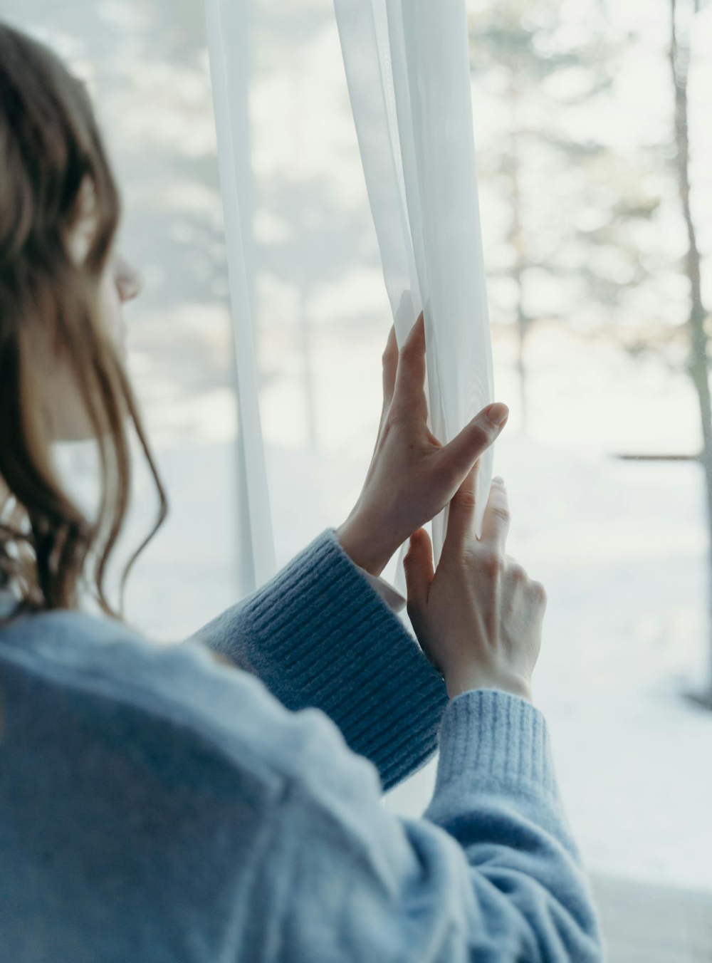 A woman wearing a blue jumper closing sheer curtains at a window.