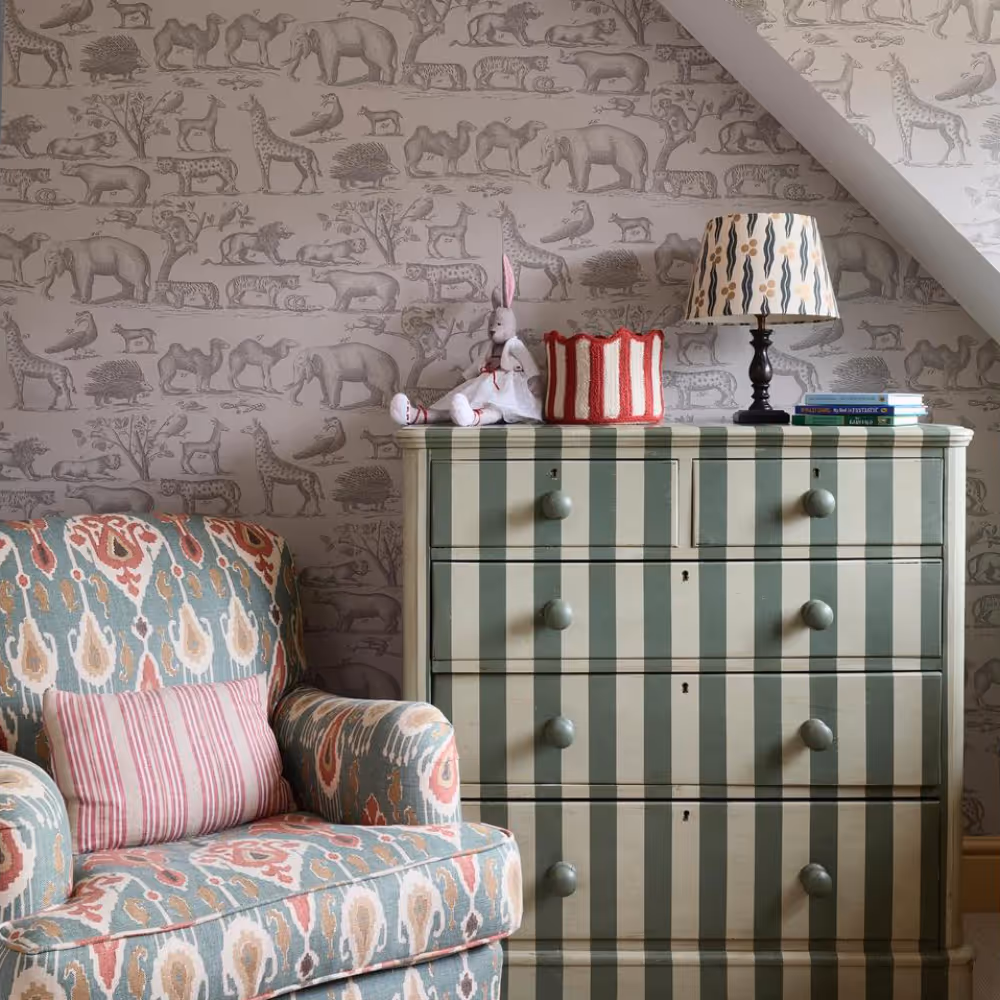 A cream and green striped chest of drawers and traditional armchair in front of a wall with an animal theme wallpaper